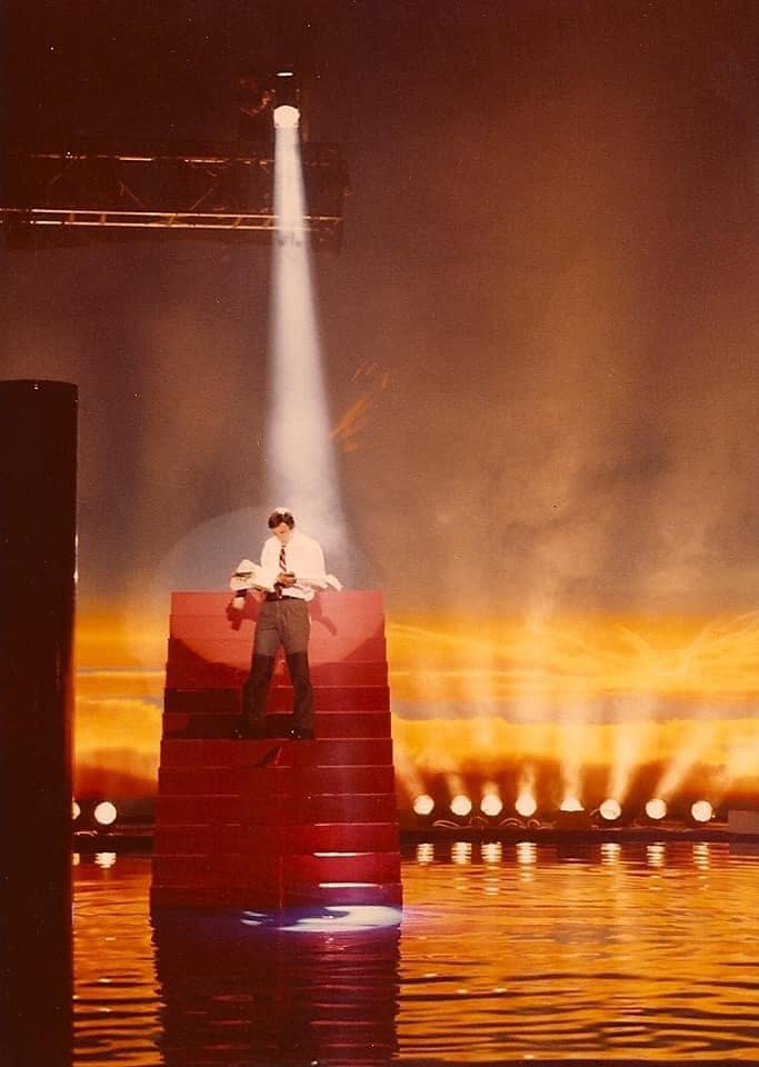 Bill Spooner atop a red stage pyramid under a single spotlight, orange fire and water backdrop
