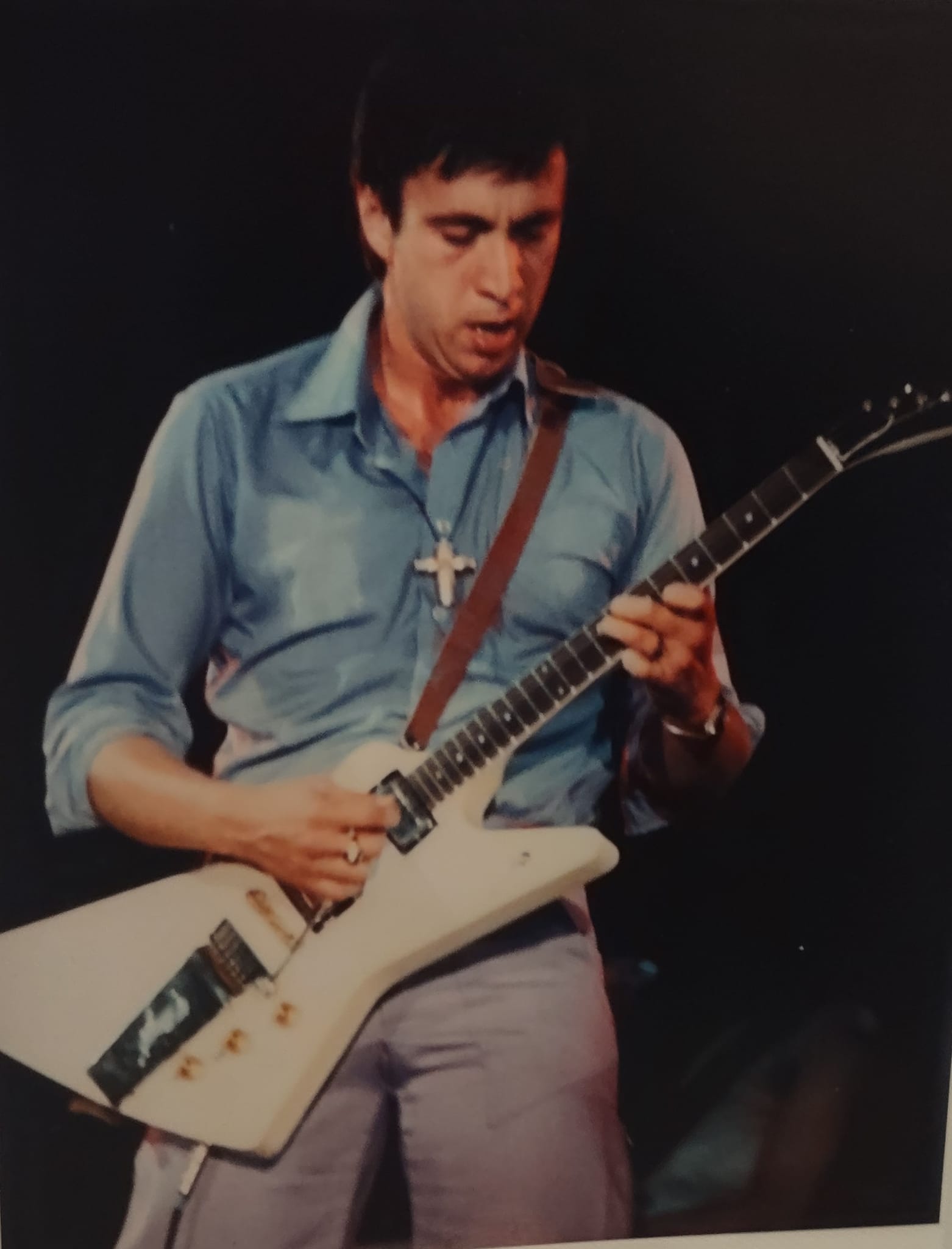 Bill Spooner in a blue shirt with a cross necklace, leaning into his white Flying V guitar on stage — intense color close-up