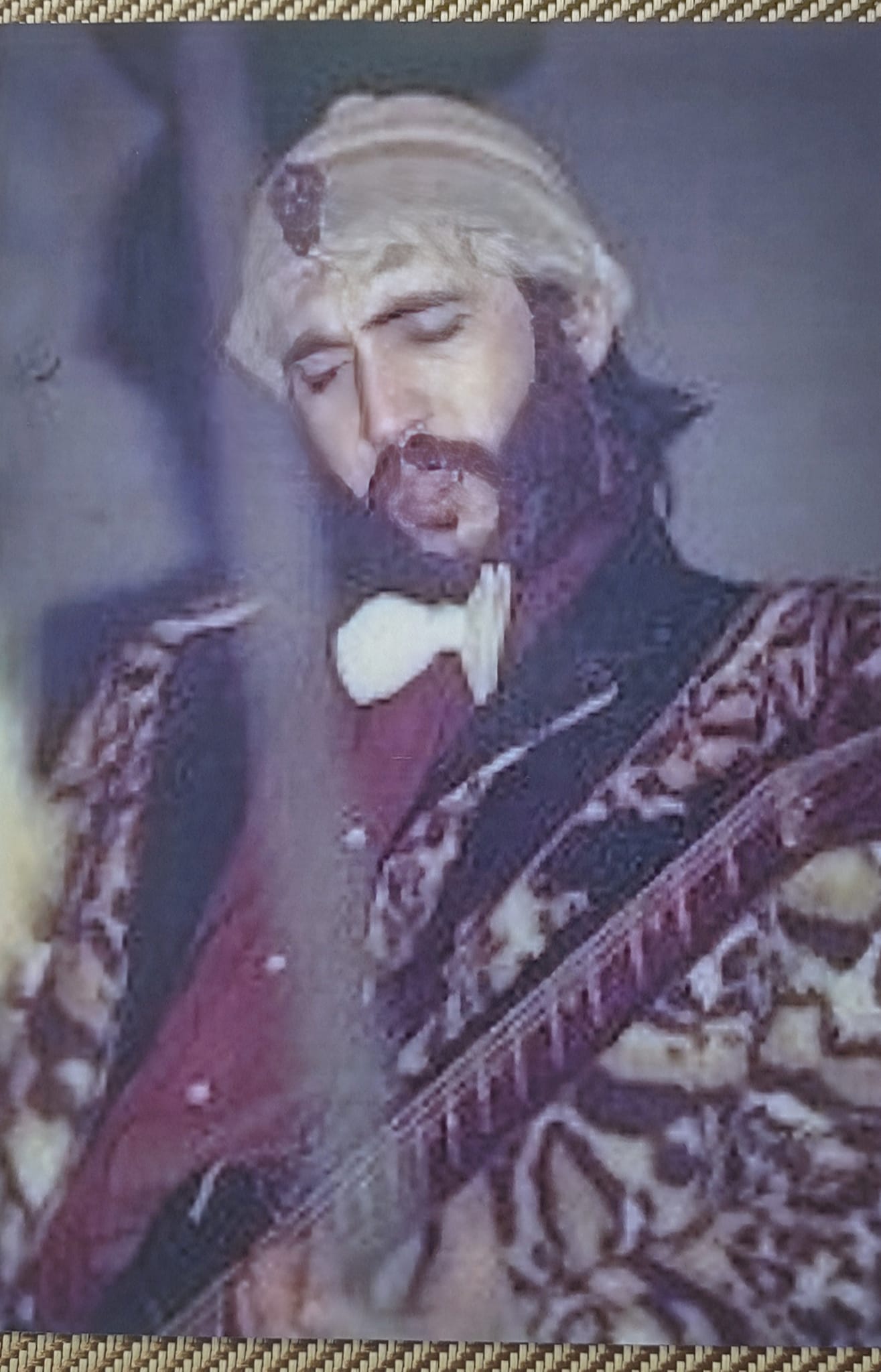 Bill Spooner in an ornate theatrical costume with blonde wig and bow tie, eyes closed, playing guitar — color stage shot