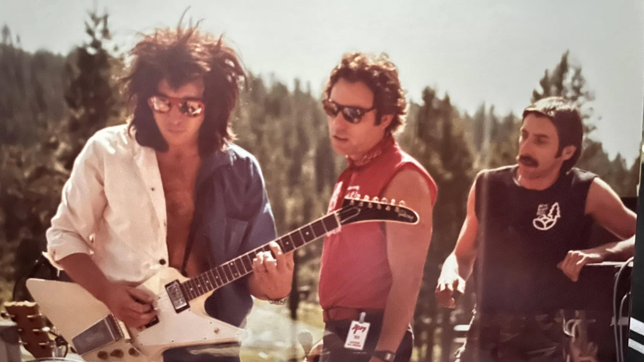 Bill Spooner in a white shirt playing his white Flying V outdoors with two companions — color candid shot
