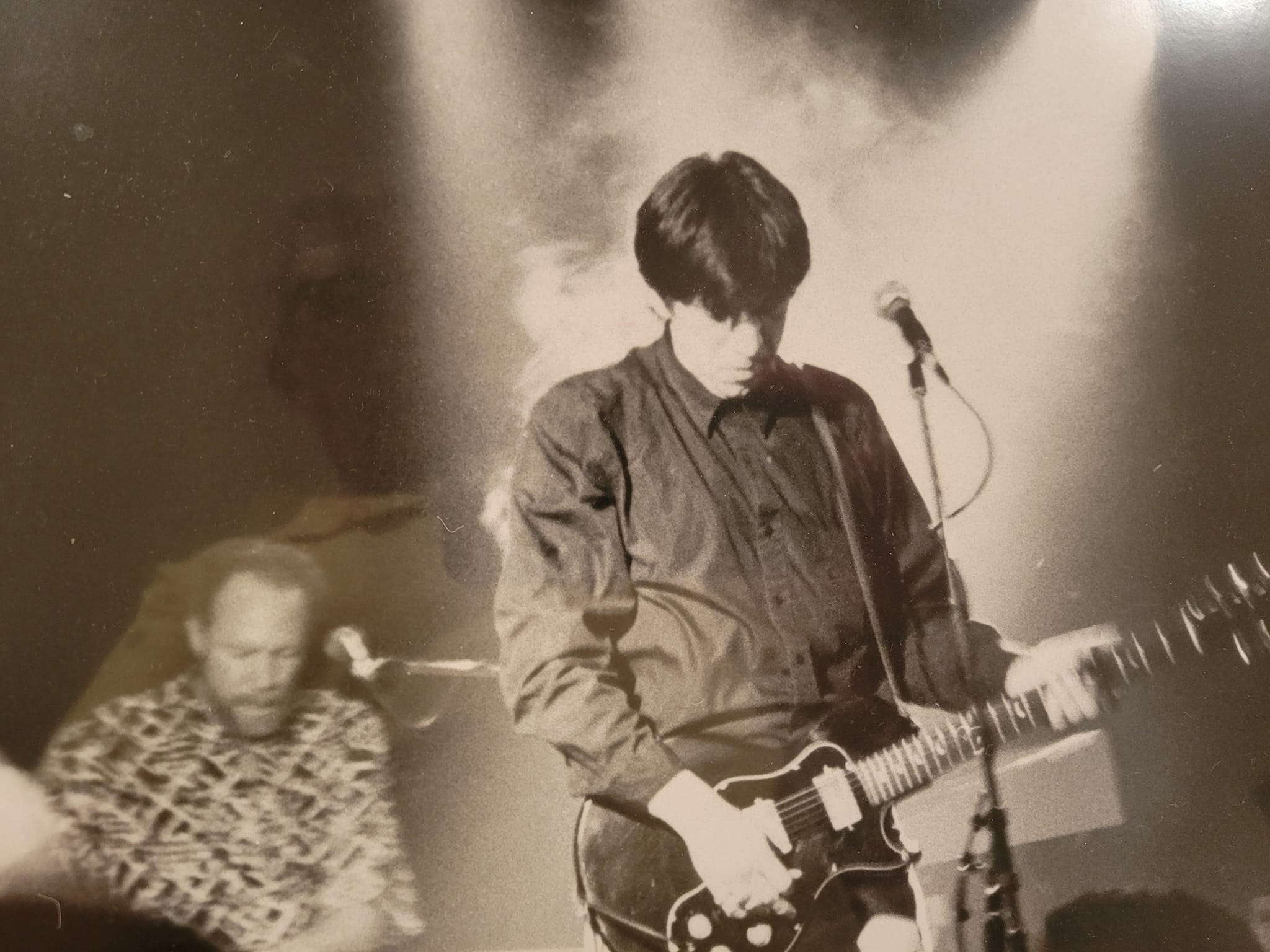 Bill Spooner on stage playing an SG-style guitar under spotlights with smoke behind him, keyboard player in background — black-and-white