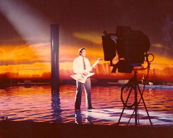 Bill Spooner playing his white Flying V on a TV stage set — sunset backdrop, camera beside him