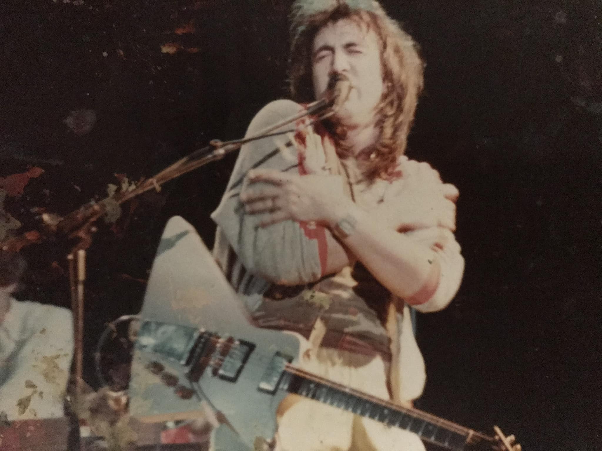 Bill Spooner arms wrapped around the mic stand on stage, long hair, intense expression — color live shot
