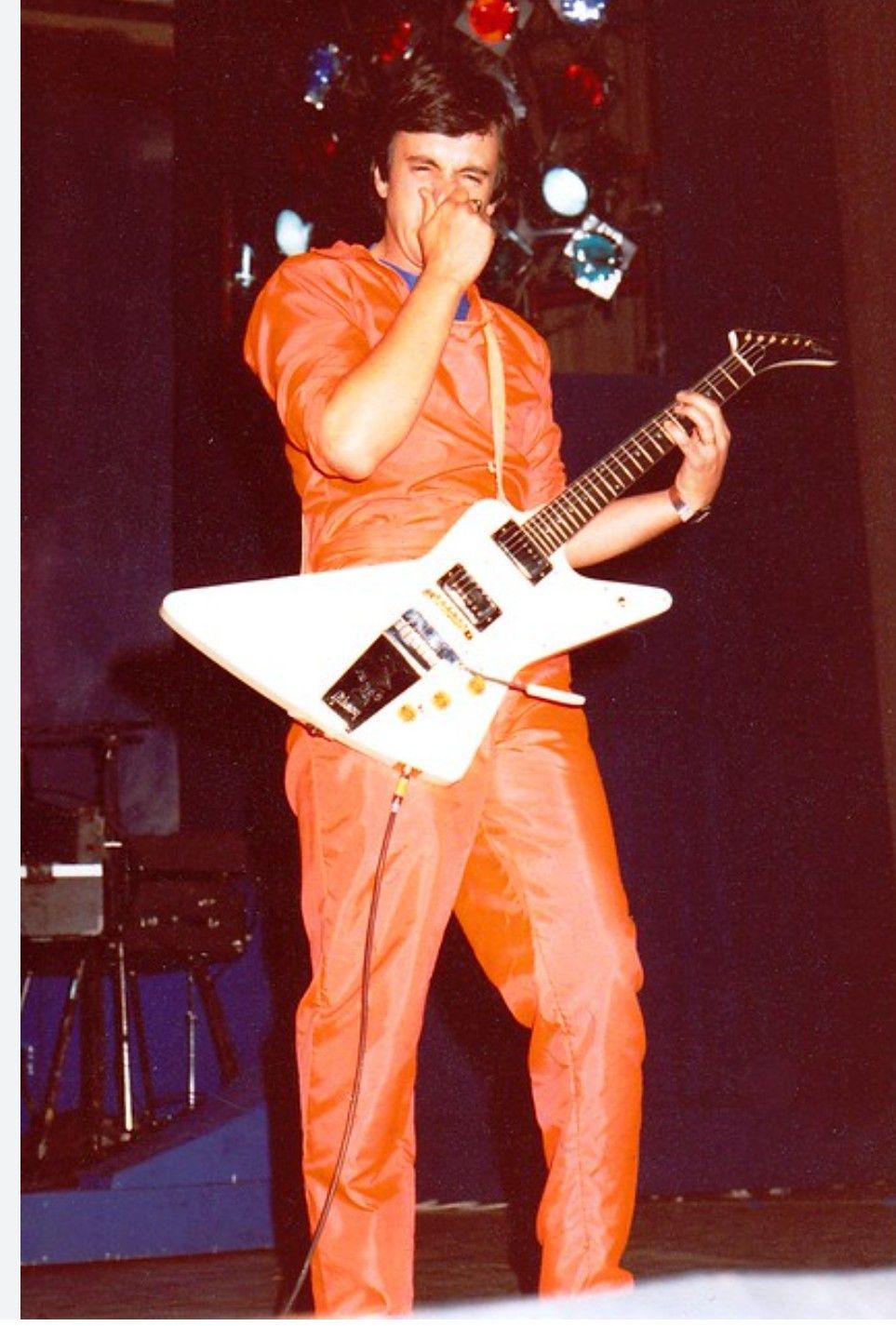 Bill Spooner on stage in an orange jumpsuit playing a white Flying V guitar