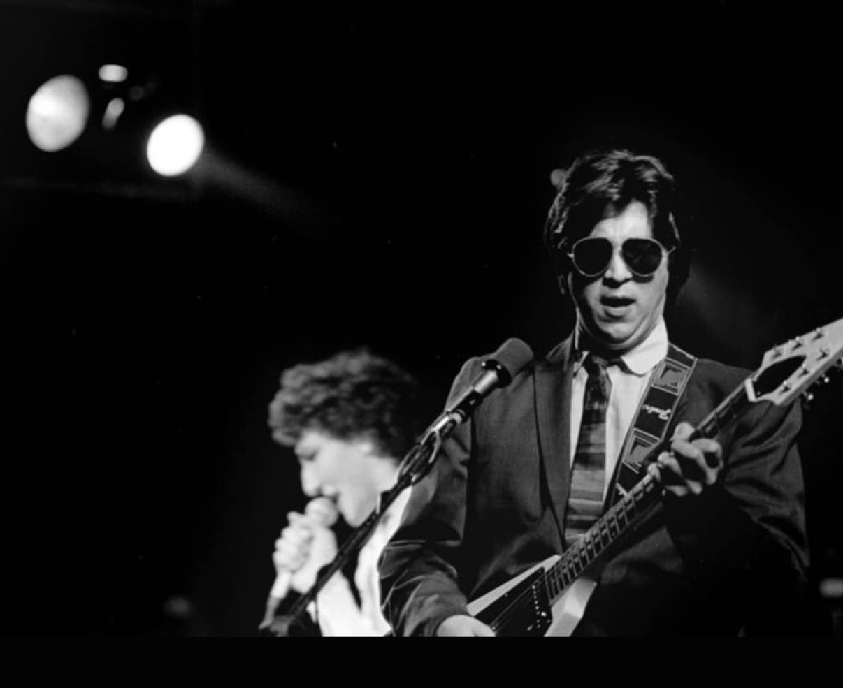 Bill Spooner in sunglasses playing guitar, singer with microphone in soft focus at left — black-and-white live shot