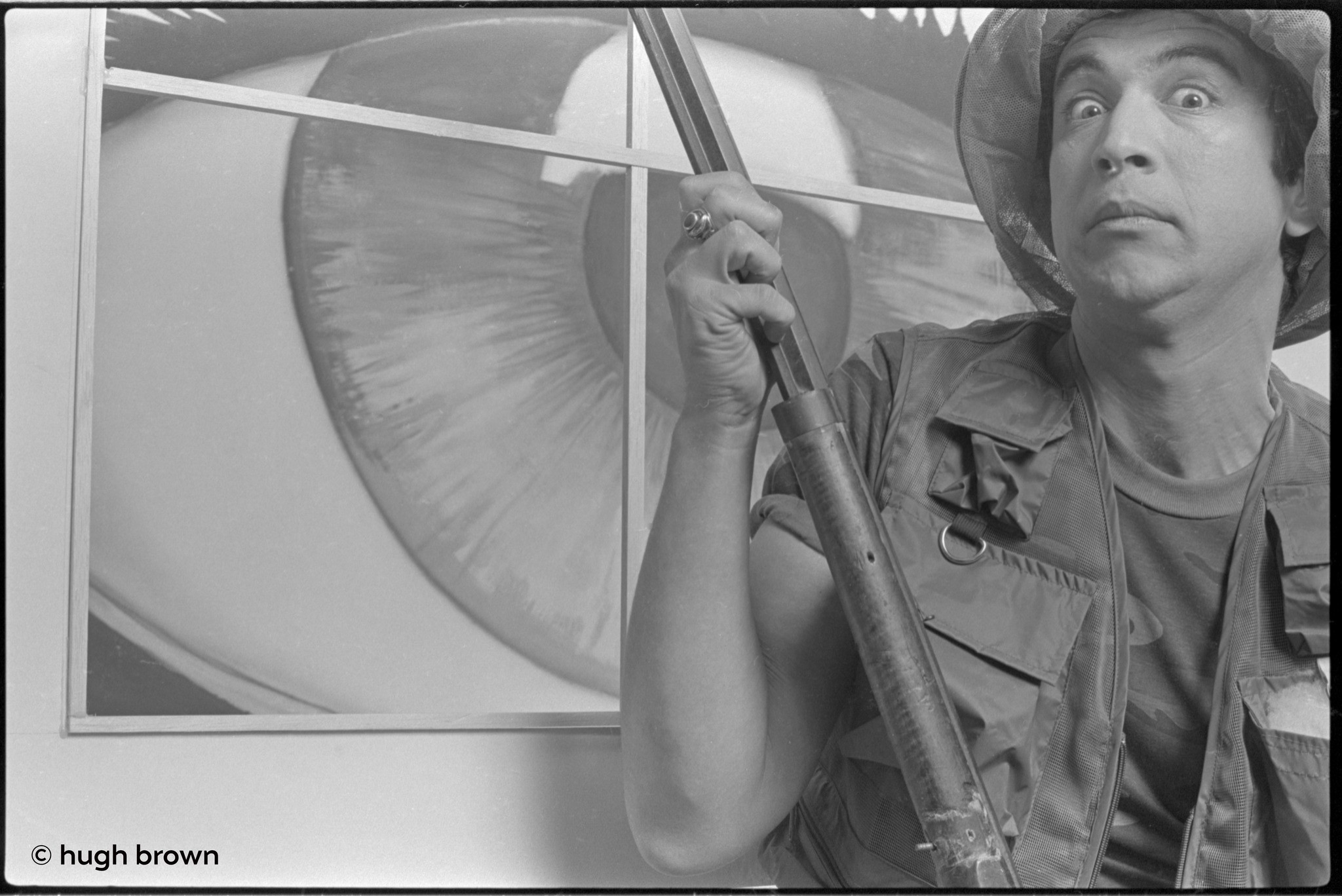Bill Spooner wide-eyed in a military safari hat with a prop rifle next to a framed eye artwork — dramatic black-and-white, photo: Hugh Brown