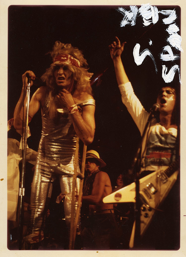 Fee Waybill as Quay Lewd in a silver jumpsuit and platform boots, with Bill Spooner on Flying V beside him