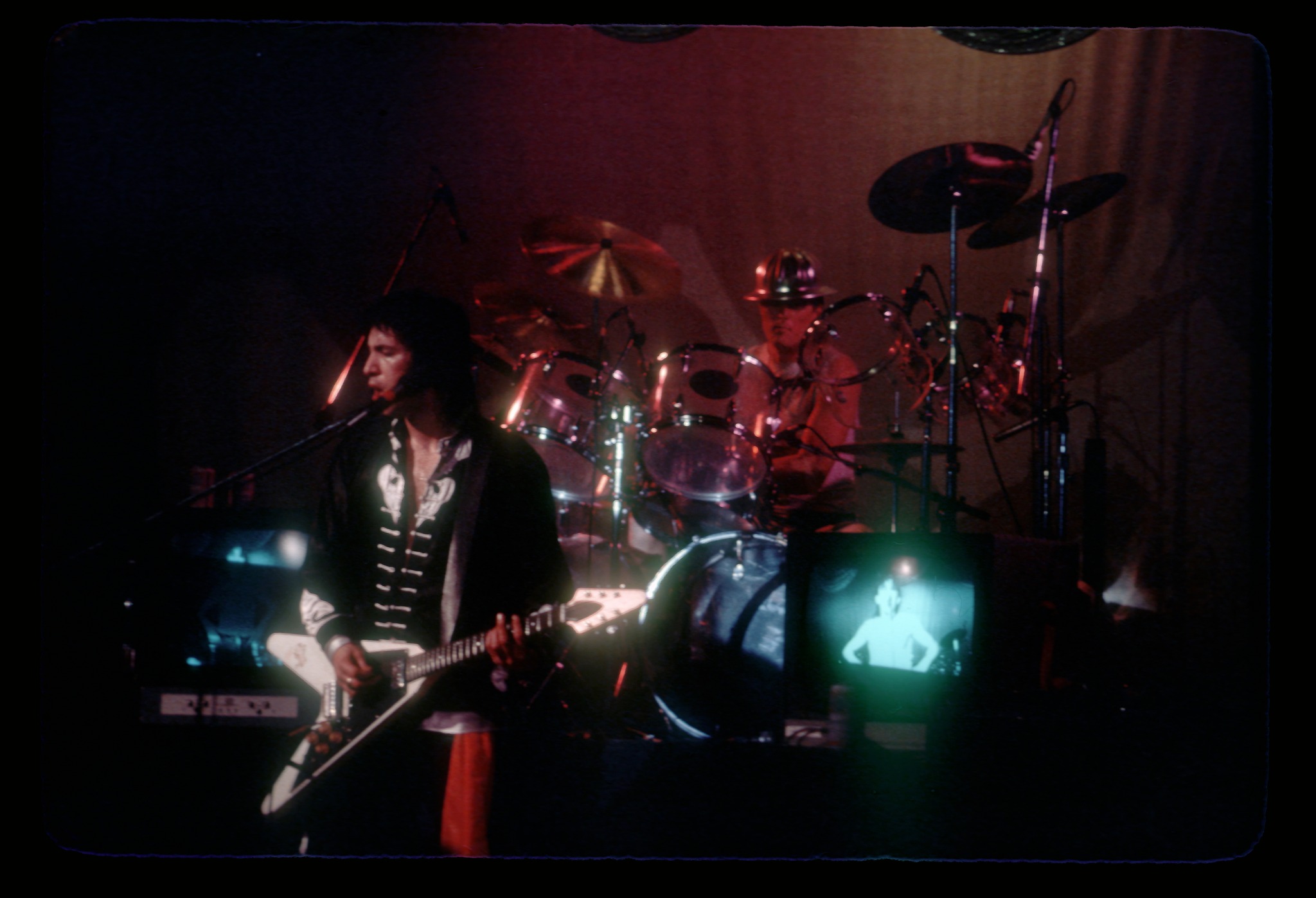 Bill Spooner on Flying V in the foreground with Prairie Prince drumming behind him, purple stage lights and a TV monitor on stage