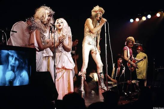 The Tubes on stage in 1973 — Fee Waybill in platform boots at the mic, costumed performers in white beside him, a television monitor at the stage edge
