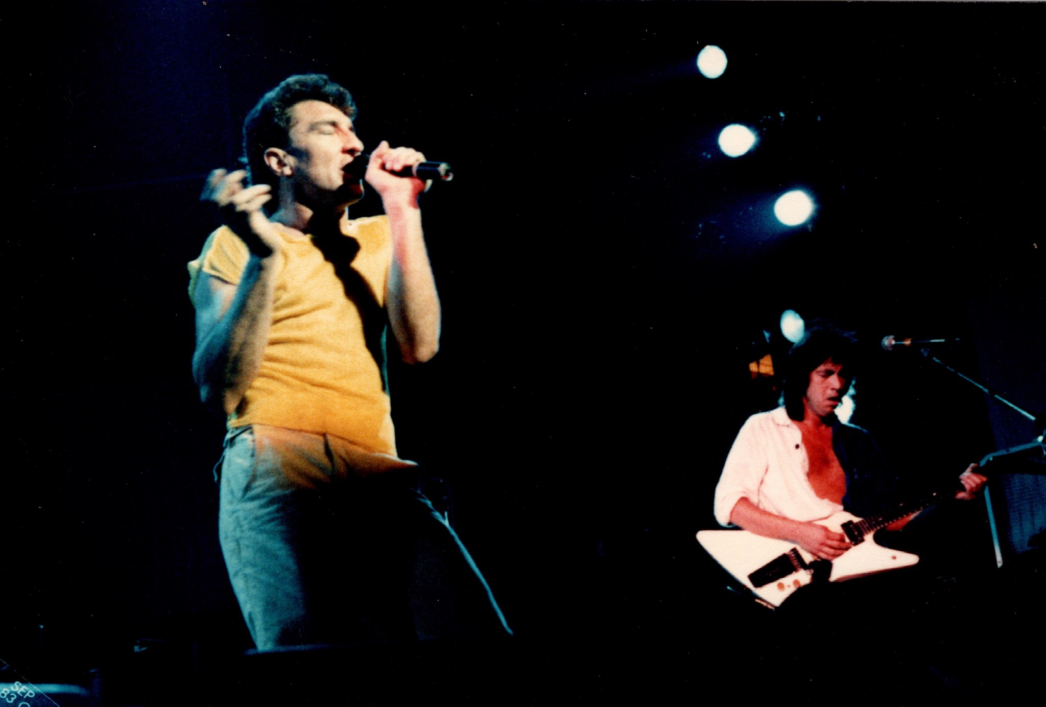 Fee Waybill singing in a yellow t-shirt with Bill Spooner on white Flying V in the background — color live shot