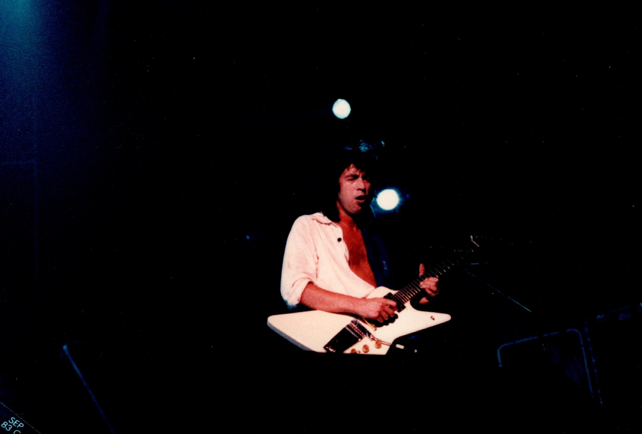 Bill Spooner alone on stage playing his white Flying V guitar under blue spotlights — color, September 1983