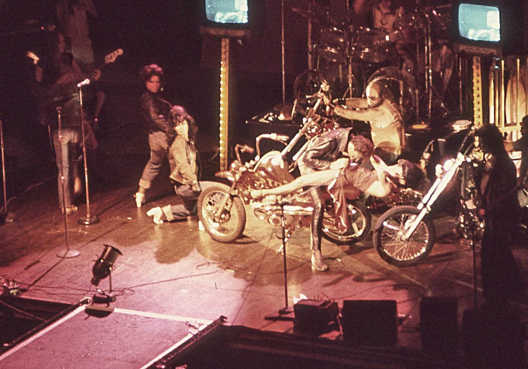 The Tubes on stage with a motorcycle prop beneath banks of televisions