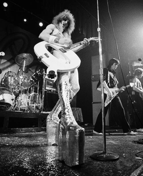 Quay Lewd in towering silver platform boots holding a Flying V guitar on stage, black and white
